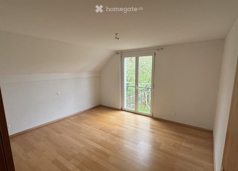 Empty attic room with wooden flooring, white walls, and a large sliding glass door leading to a balcony.