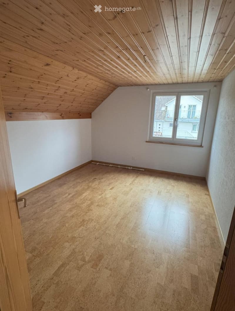 Empty room with wooden floor and slanted wooden ceiling, featuring a single window with white frame and a door with brass handle on the left side.