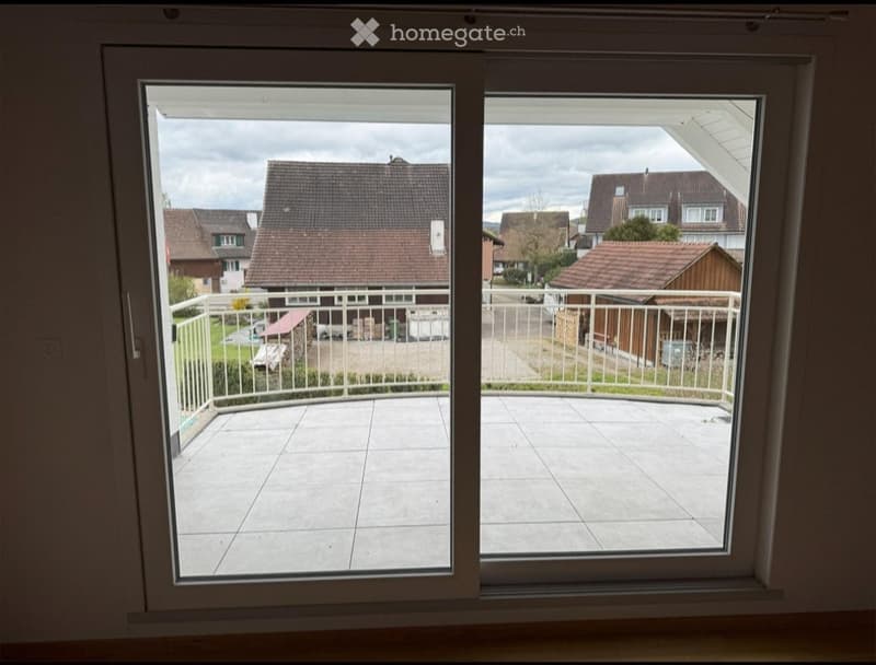 Balcony with tiled floor, white metal railing, overlooking a residential area with houses and greenery.