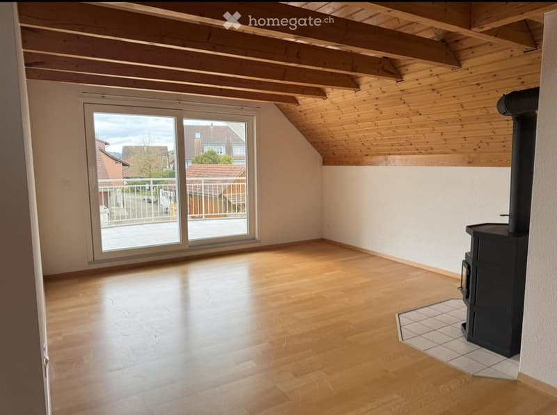 Empty attic room with wooden floors, exposed wooden beams, large window, and a fireplace.