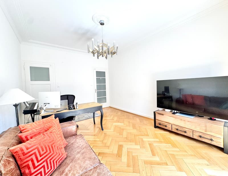 A modern living room with a wooden floor, a large sofa with red pillows, a TV on a wooden stand, a desk with a laptop, and a chandelier hanging from the ceiling.