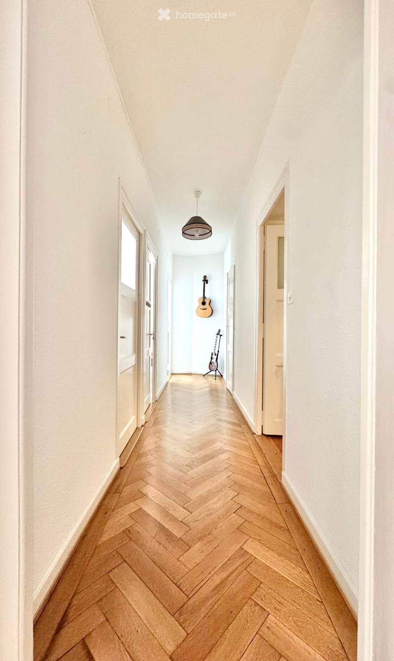 A long, narrow hallway with herringbone-patterned wooden flooring, white walls, and a hanging pendant light. Two guitars are displayed on stands against the far wall.