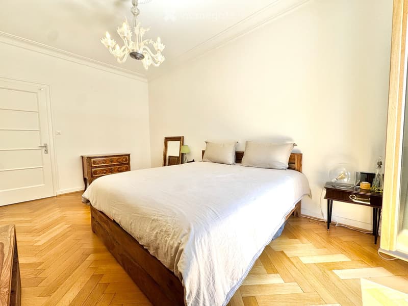 Elegant bedroom with a wooden bed, nightstand, mirror, and chandelier. The room features light-colored bedding and wooden flooring.