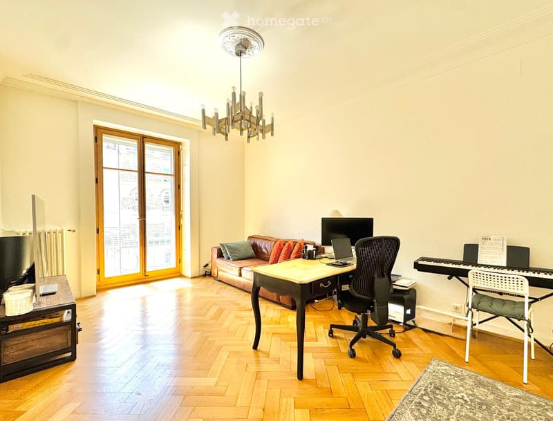 Bright living room with wooden floors, large windows, a sofa, a desk with a laptop, a piano, and a chandelier.