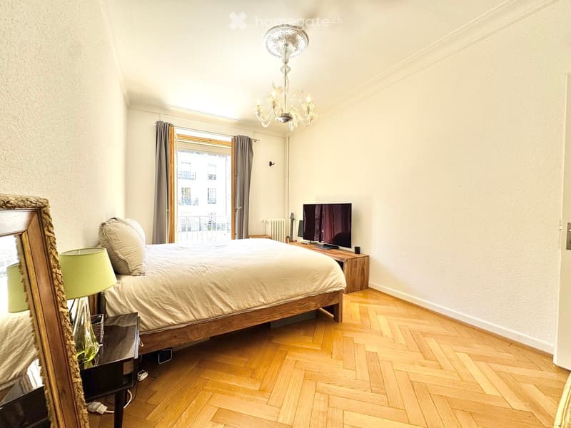 Cozy bedroom with wooden herringbone flooring, a large bed with white bedding, a nightstand with a lamp, a mirror, and a television on a wooden stand. A guitar is placed near the window, and the room features a chandelier and large windows with curtains.