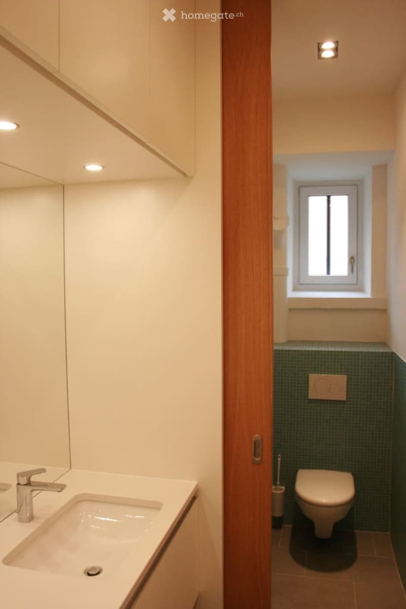 Modern bathroom with a white sink, chrome faucet, mirror, and a toilet. The walls are painted in a light color, and there is a small window providing natural light.