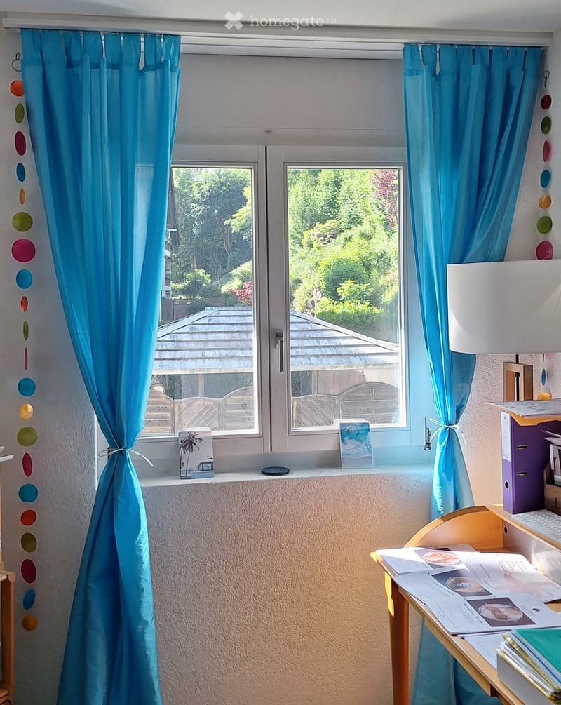 A well-lit room with a desk, a lamp, and colorful curtains. The window offers a view of a garden and a wooden deck.