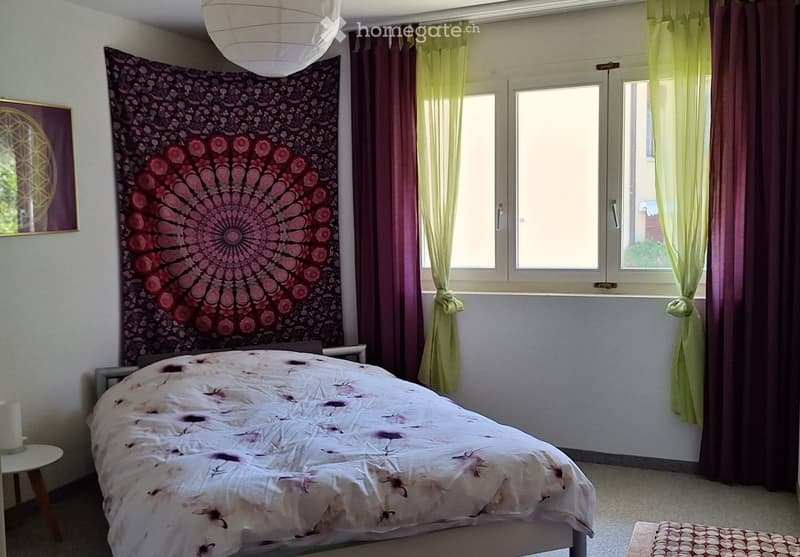 Cozy bedroom with a double bed, nightstand, small stool, colorful tapestry, and large windows with sheer curtains.