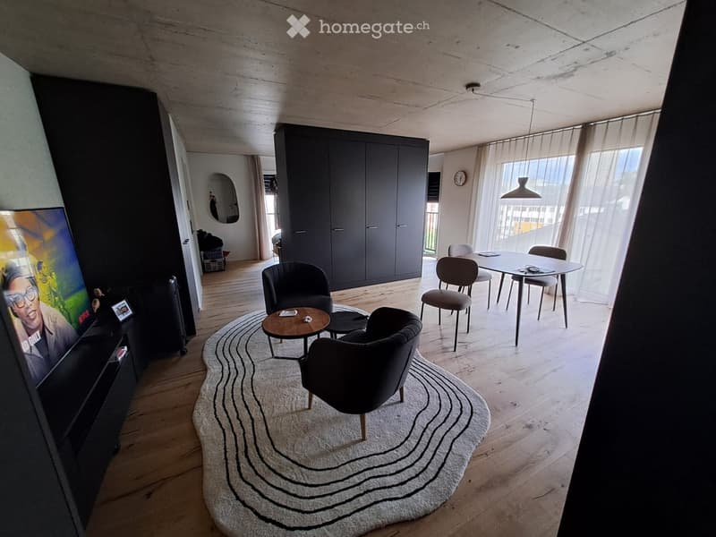 Modern living room with wooden flooring, a large round rug, a coffee table, chairs, a dining table, and a TV on a stand.
