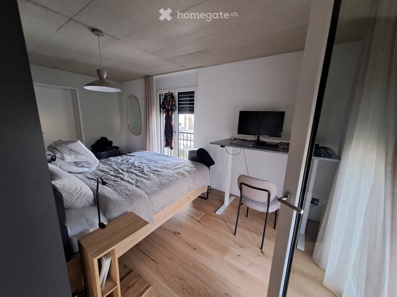 A modern, compact bedroom with a bed, a desk with a screen, a chair, a small wooden shelf, and a mirror. The room features light wooden flooring and white walls.