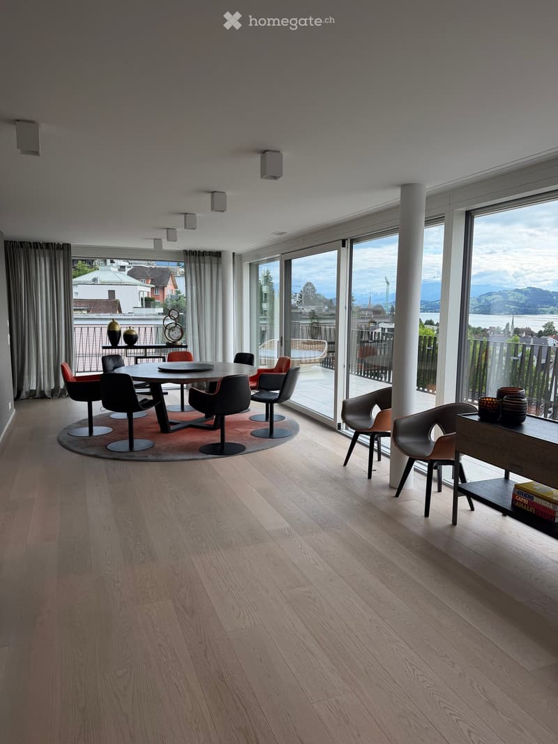Modern dining area with wooden floor, large windows offering a scenic view, dining table with chairs, decorative vases, and a shelf with books.