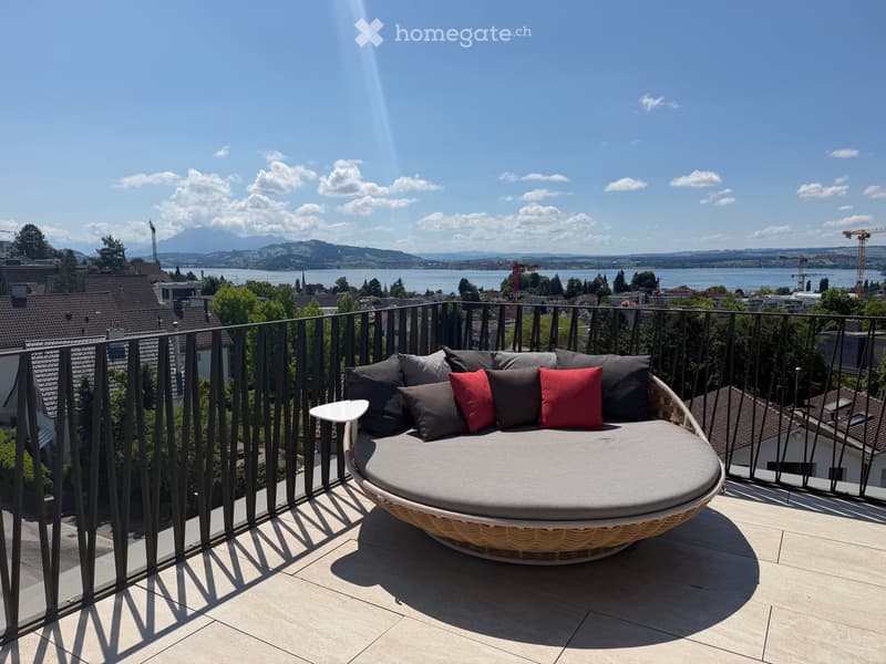 Balcony with a round wicker sofa, red and grey cushions, and a small white table, overlooking a scenic lake and mountains.