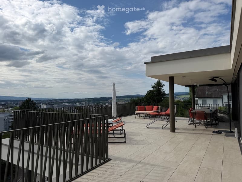 Balcony with tiled floor, metal railing, lounge chairs, sofa, table, and umbrella, overlooking a cityscape with trees and hills.