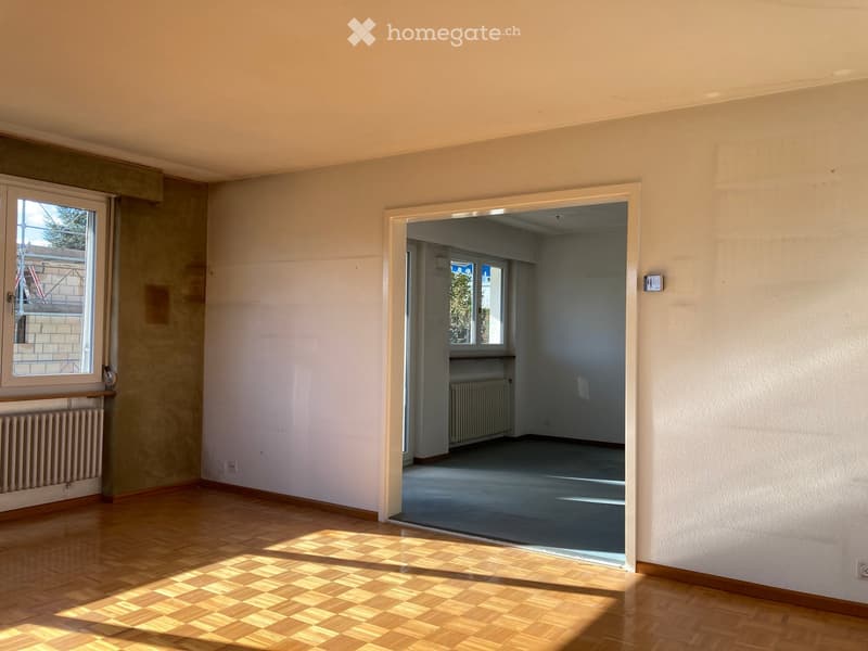 An empty room with two windows, radiators, wooden floor, a door that leads to another room