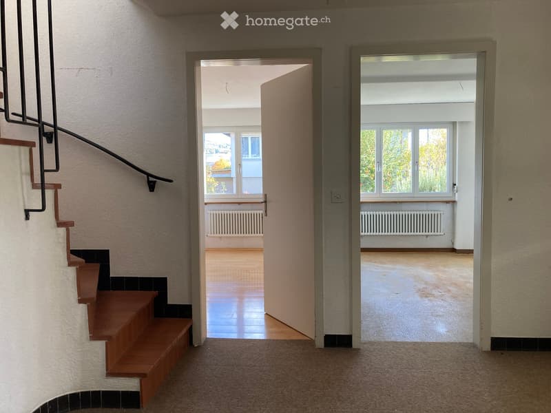 Spacious hallway with wooden staircase, open doors leading to two rooms with large windows and radiators.