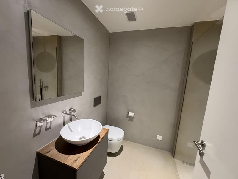 Modern bathroom with a sleek, minimalist design. Features include a white sink with a modern faucet, a large mirror, a toilet, and a wooden cabinet.