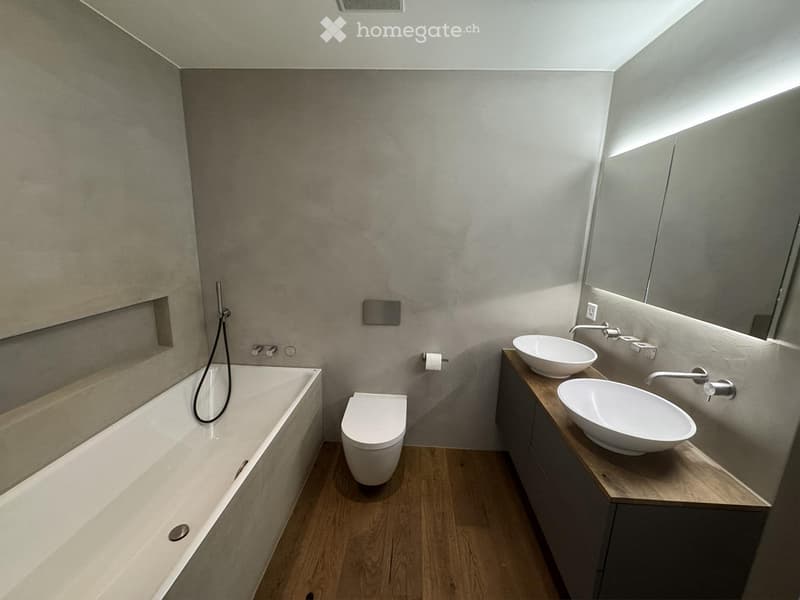 Modern bathroom with a bathtub, toilet, double sink, and large mirror. The walls are grey, and the floor is wooden.