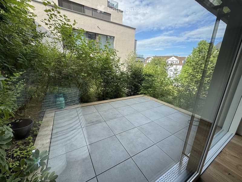 Balcony with tiled floor, glass railing, and view of greenery and buildings.