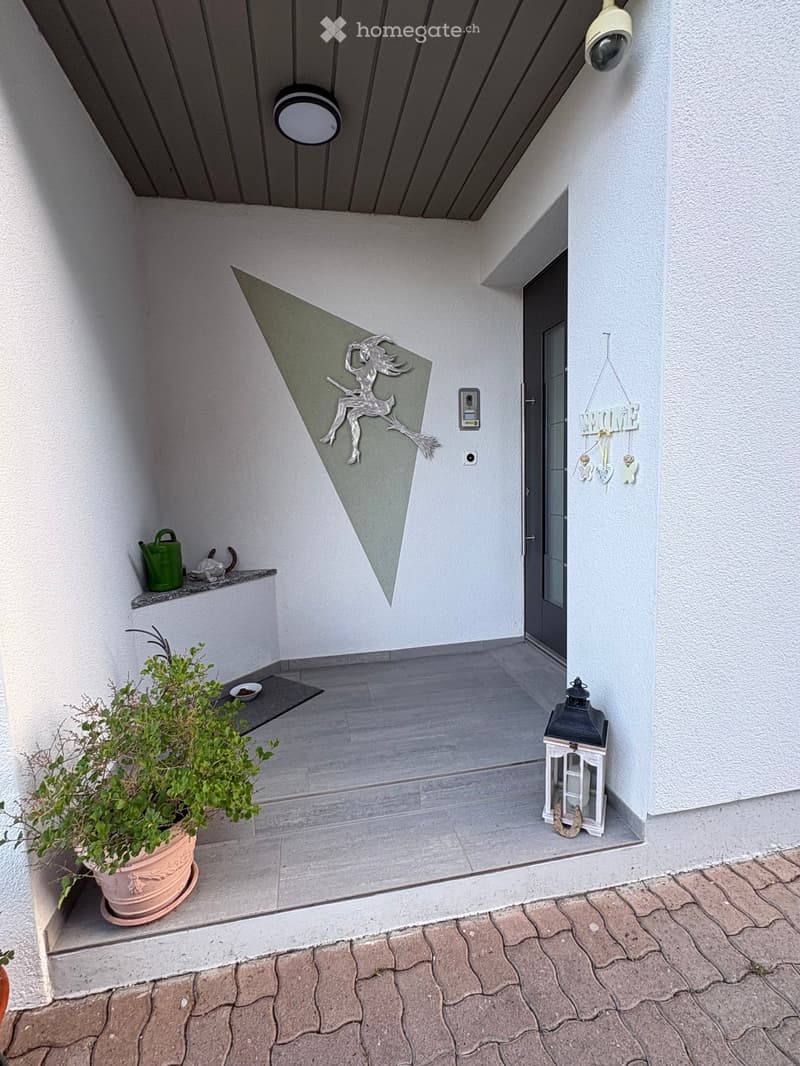 Entrance area with a decorative metal sculpture, lantern, potted plant, and a small shelf with a watering can and cat figurine.