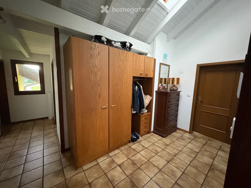 A small, furnished room with tiled flooring, a large wooden wardrobe, a mirror, a lamp, a small drawer unit, and some clothing and shoes placed on the floor.