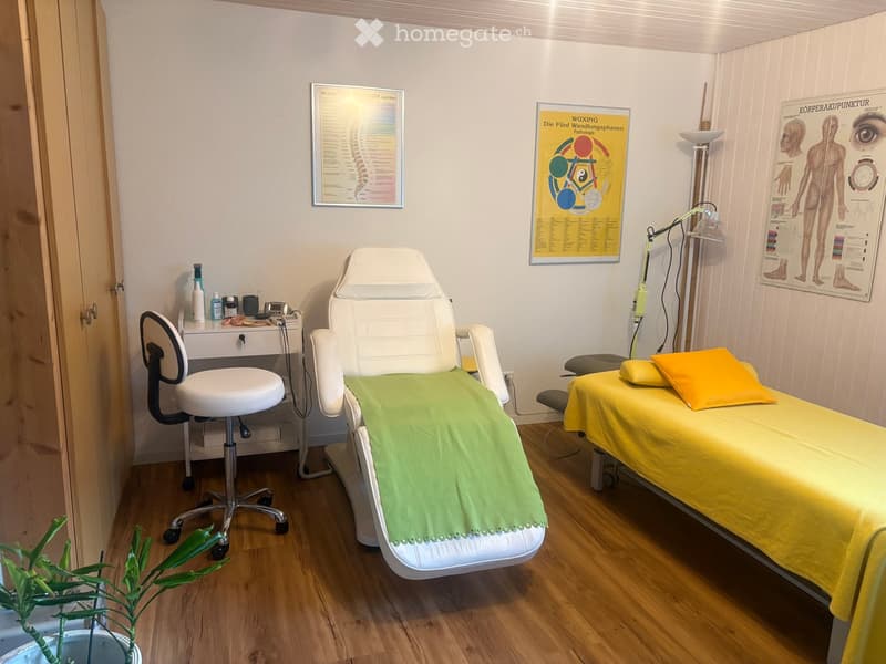 A well-lit room with wooden flooring, two treatment tables covered in green and yellow sheets, a white chair, a small table with various items, and posters on the walls.