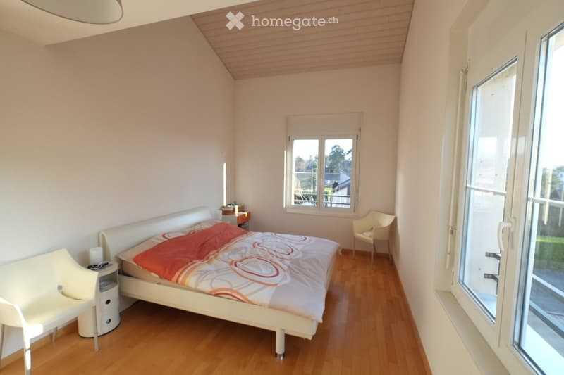 Bright bedroom with wooden flooring, white walls, and a slanted wooden ceiling. Features a white bed with a colorful duvet, a nightstand with a lamp, and a white chair near a large window with a view.