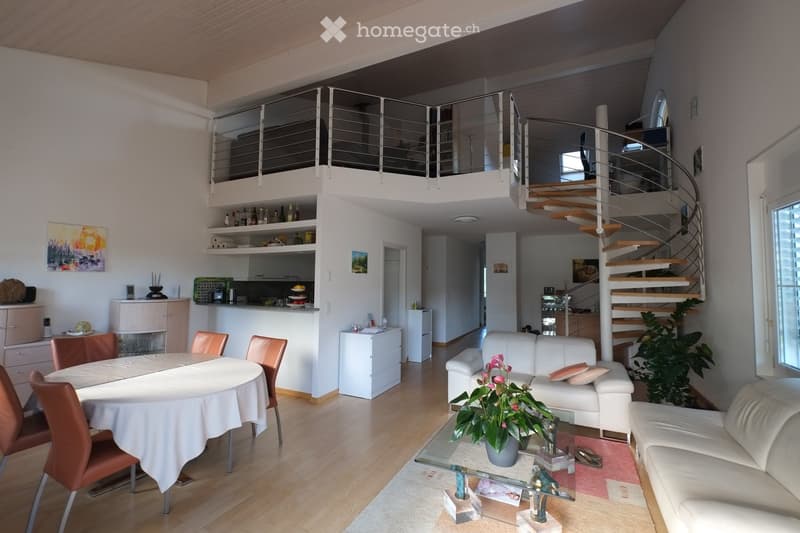 Open-plan living and dining area with white walls, wooden flooring, a white sofa, glass coffee table, dining table with chairs, kitchen area, and a spiral staircase leading to a lofted area.