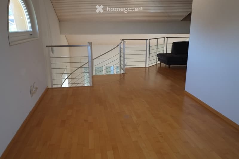 A modern, open-plan living space with wooden flooring, white walls, and a curved staircase with a metal railing. A single black sofa is positioned near the staircase, and a small window is visible on the left side.