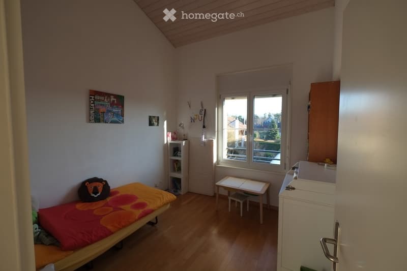 Cozy bedroom with wooden floor, slanted ceiling, bed with colorful blanket, small table, stool, shelf, and window with view of greenery.