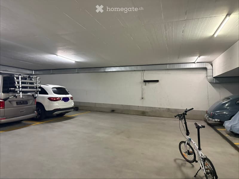 Underground parking area with several parked cars, including a white SUV with a roof rack, a grey car, and a bicycle. The space features concrete floors, white walls, and fluorescent lighting.