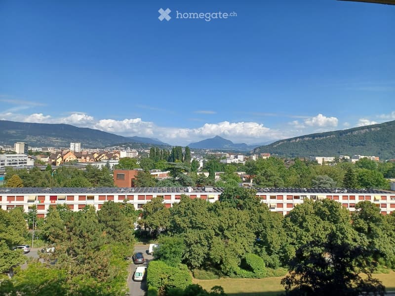 A multi-story residential building with red and white facade, surrounded by trees and parked cars, with a cityscape and mountains in the background.