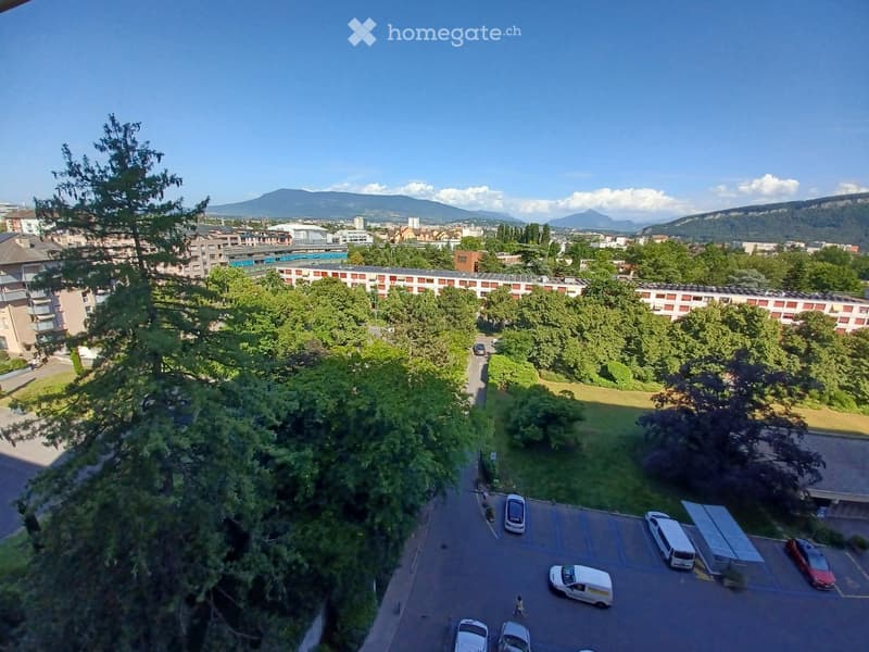 Aerial view of a residential area with a parking lot, trees, and distant mountains under a clear blue sky.