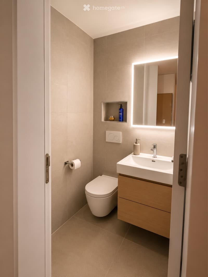 Modern bathroom with beige tiles, a floating wooden vanity with a sink, a toilet, a large illuminated mirror, and a small shelf holding toiletries.