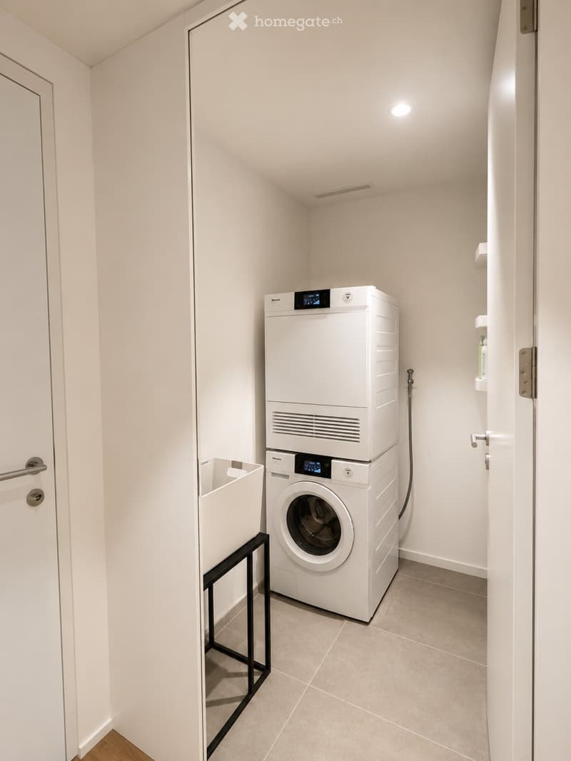 Compact bathroom with a stacked washing machine and dryer unit, white walls, tiled floor, and a small shelf with cleaning supplies.