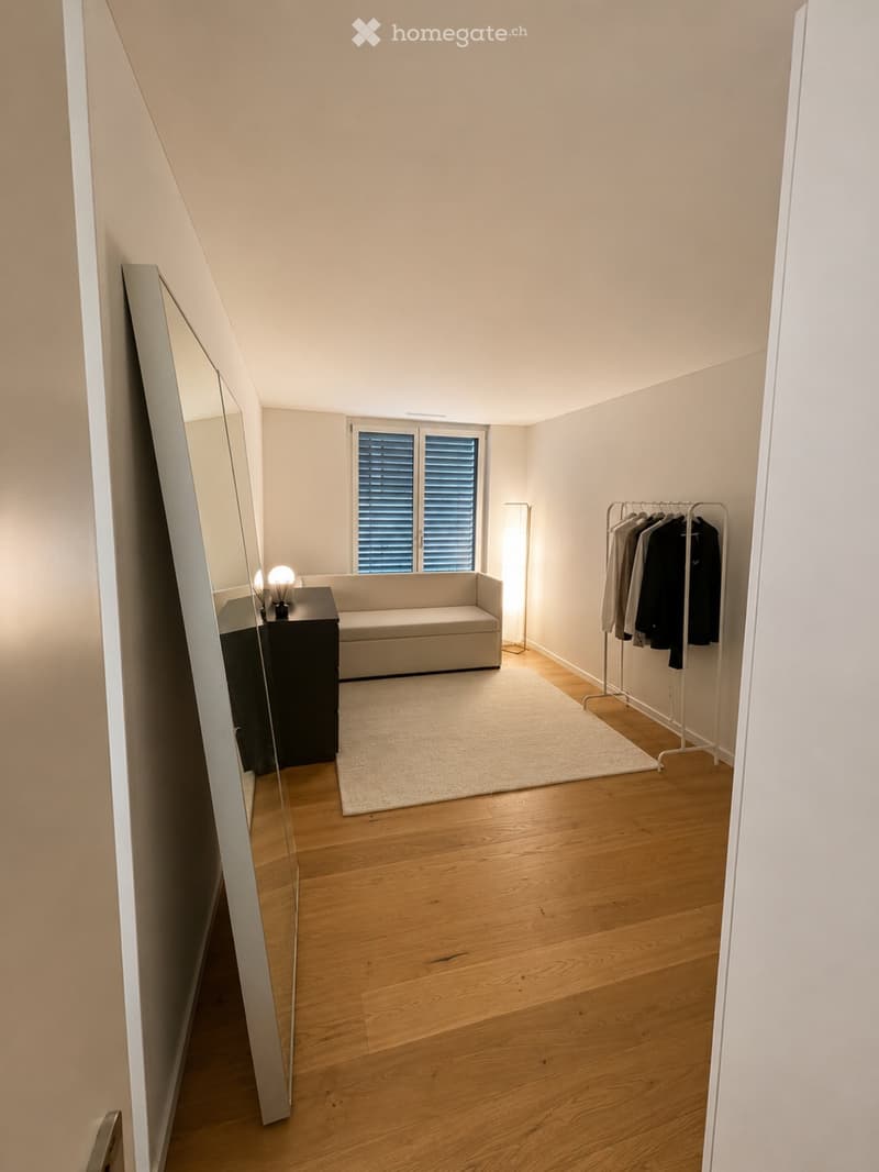 A modern, minimalist living room with light wooden flooring, a white sofa, a small table with a lamp, a clothing rack, and a large window with blinds.