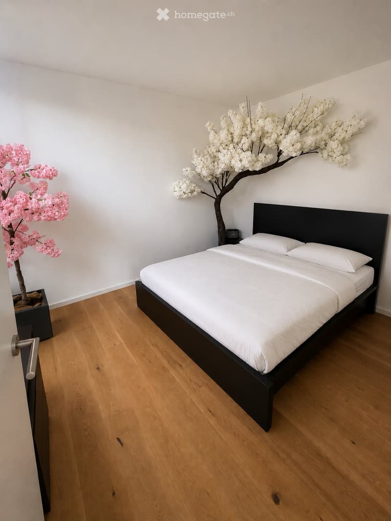 Modern bedroom with a large bed, white bedding, and two decorative artificial trees in pots. The room features light wooden flooring and white walls.