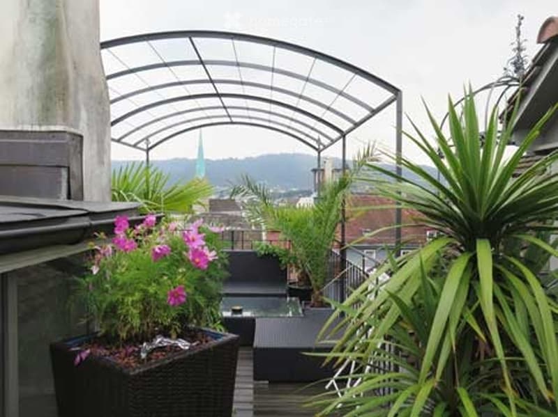 Rooftop terrace with a metal pergola, potted plants, and a view of the city and mountains.