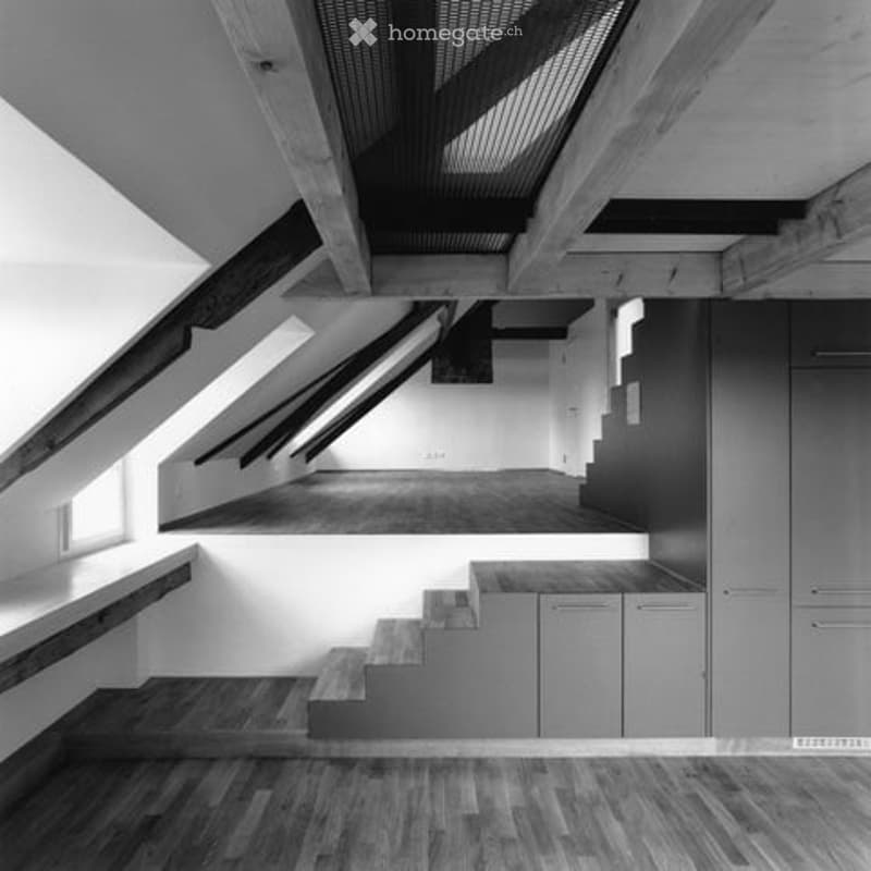 Modern, minimalist kitchen with sleek cabinets and a wooden staircase leading to an upper level. The space features exposed wooden beams and ample natural light from skylights.