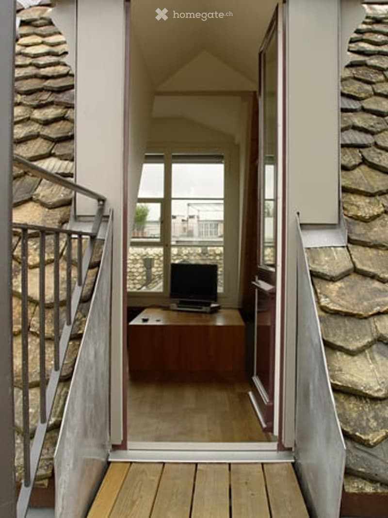 A small, modern room with wooden flooring, a table with a laptop and screen, and a staircase leading upwards.