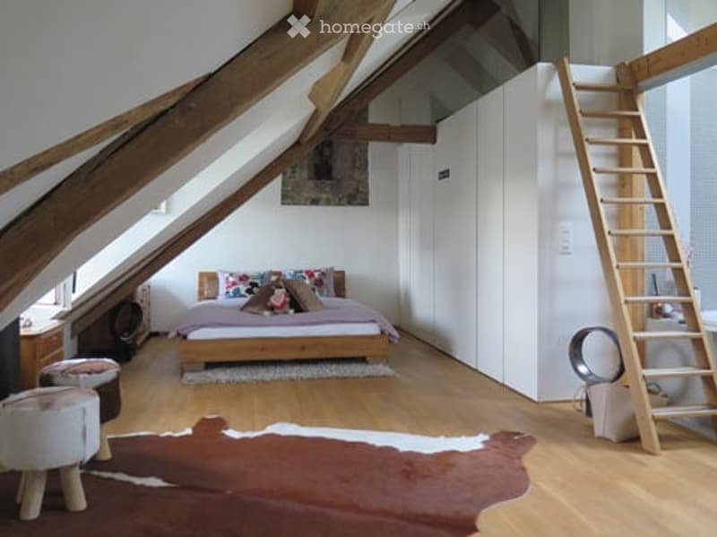 Cozy attic bedroom with wooden beams, slanted ceilings, and large windows. Features a bed with colorful pillows, a nightstand, a cowhide rug, a wardrobe, and a wooden ladder leading to a loft area.