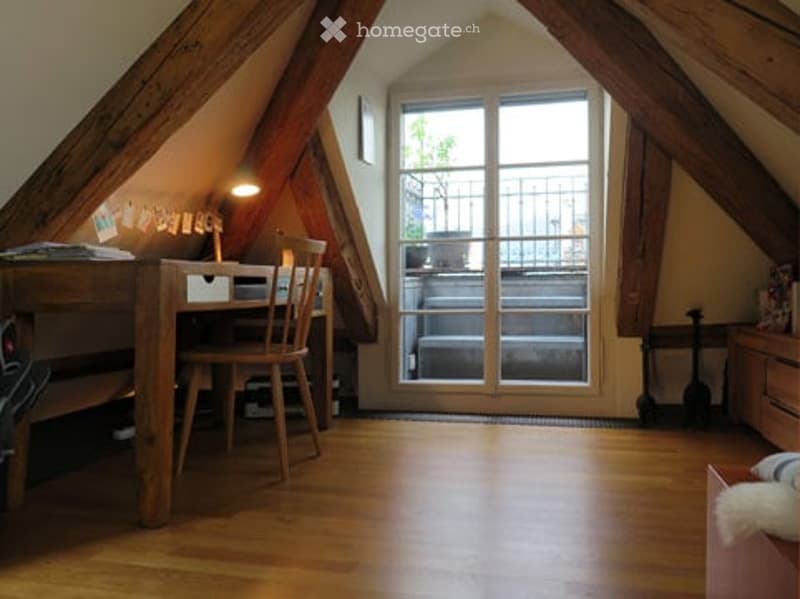 Cozy attic room with wooden floors, slanted wooden beams, large window with balcony access, desk, chair, and lamp.
