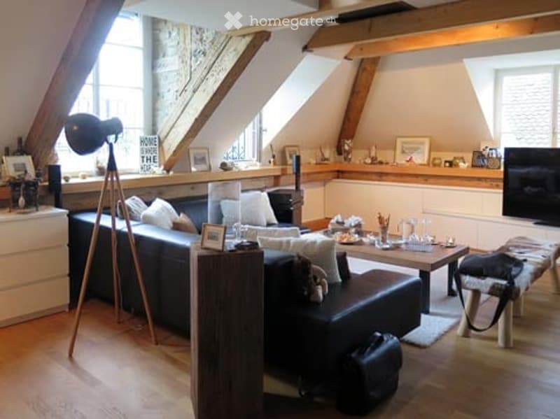 Cozy attic living room with exposed wooden beams, large windows, dark leather sofa, coffee table, ottoman, floor lamp, TV, and decorative picture frames on the wall.