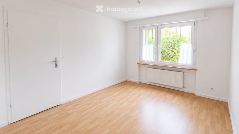 Empty room with light wooden flooring, white walls, a white door, and a window with white frames and curtains. A white radiator is placed below the window.