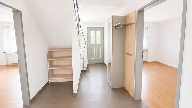 Empty hallway with wooden flooring, white walls, and a green door at the end. Shelves and a small staircase are visible on the left side.
