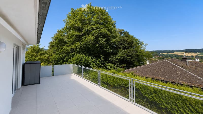 Balcony with white tiled floor, glass railing, and a view of greenery and a distant village.
