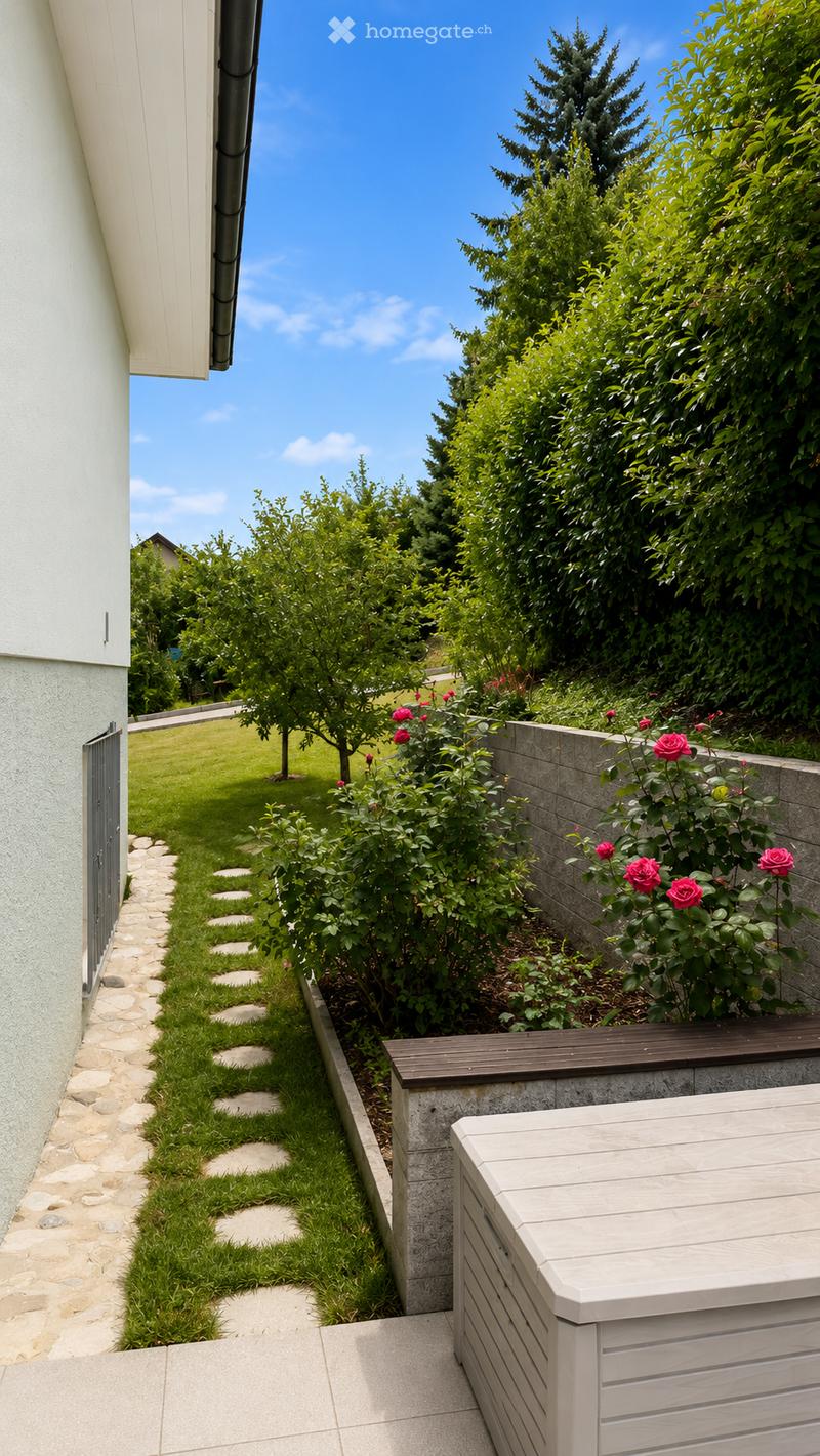 A well-maintained garden area with a stone pathway, neatly trimmed grass, and a variety of flowering plants and trees. A low concrete wall borders the garden, and a modern storage unit is visible on the right side.