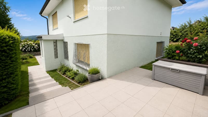 A modern garden area with a tiled patio, a grey storage bench, potted plants, and neatly trimmed hedges. The house features light-colored walls and large windows with blinds.