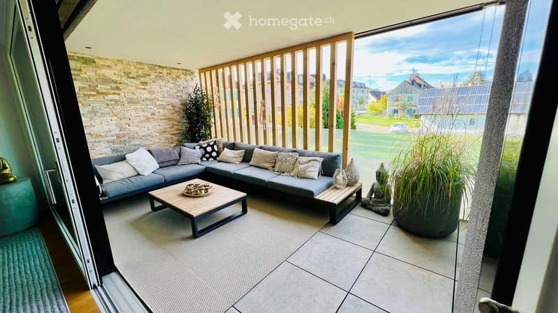 Modern, furnished balcony with a grey sectional sofa, coffee table, decorative vases, a small statue, and a large potted plant. The balcony features a stone wall, large windows with a view of the neighborhood, and a wooden privacy screen.