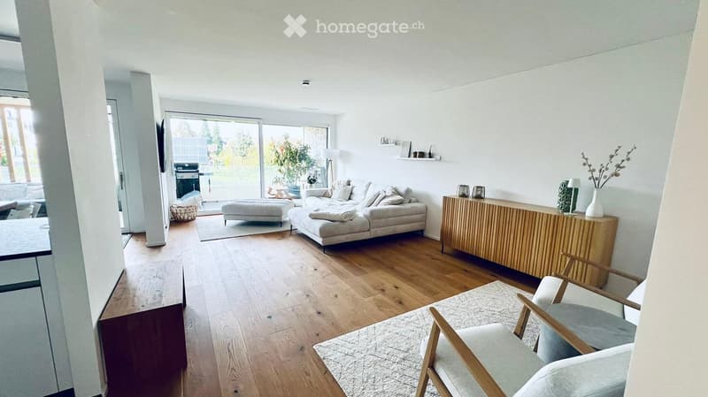 Modern living room with light wooden flooring, white walls, and large windows. Features a grey sofa with an ottoman, a wooden side table, a grey armchair, and decorative items on shelves.