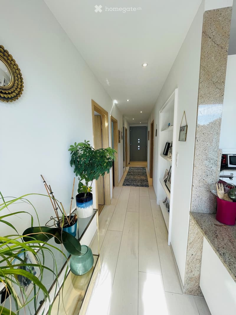 Bright hallway with white walls, wooden doors, built-in shelves, and potted plants on a glass table. The floor is tiled, and the ceiling has recessed lighting.